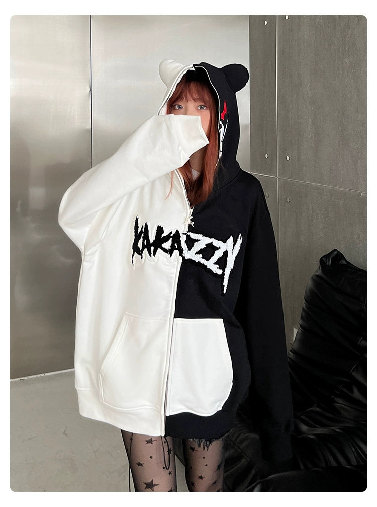 Indoors, someone wears a Seakoff Monokuma-Inspired Hoodie with ears on the hood, ideal for Danganronpa fans. The oversized black and white sweatshirt partially covers their face with one sleeve, paired with patterned leggings.