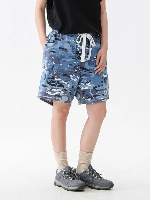 Model showcases Men's Blue Camo Drawstring Shorts - Relaxed Fit Cotton Camouflage Shorts against a plain background, styled with a black shirt, beige socks, and gray sneakers. Head and upper arms are not visible to emphasize the shorts’ style.