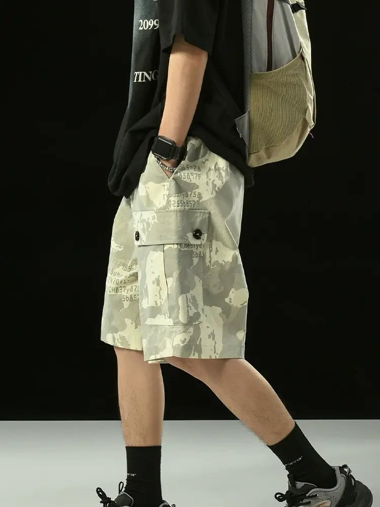 A person in a black T-shirt, Men's Grey Camo Cargo Shorts with Button Flap Pockets and Elastic Waist, black socks, sneakers, a smartwatch, and a beige backpack stands against a plain black background. Only the lower half of their face is visible.