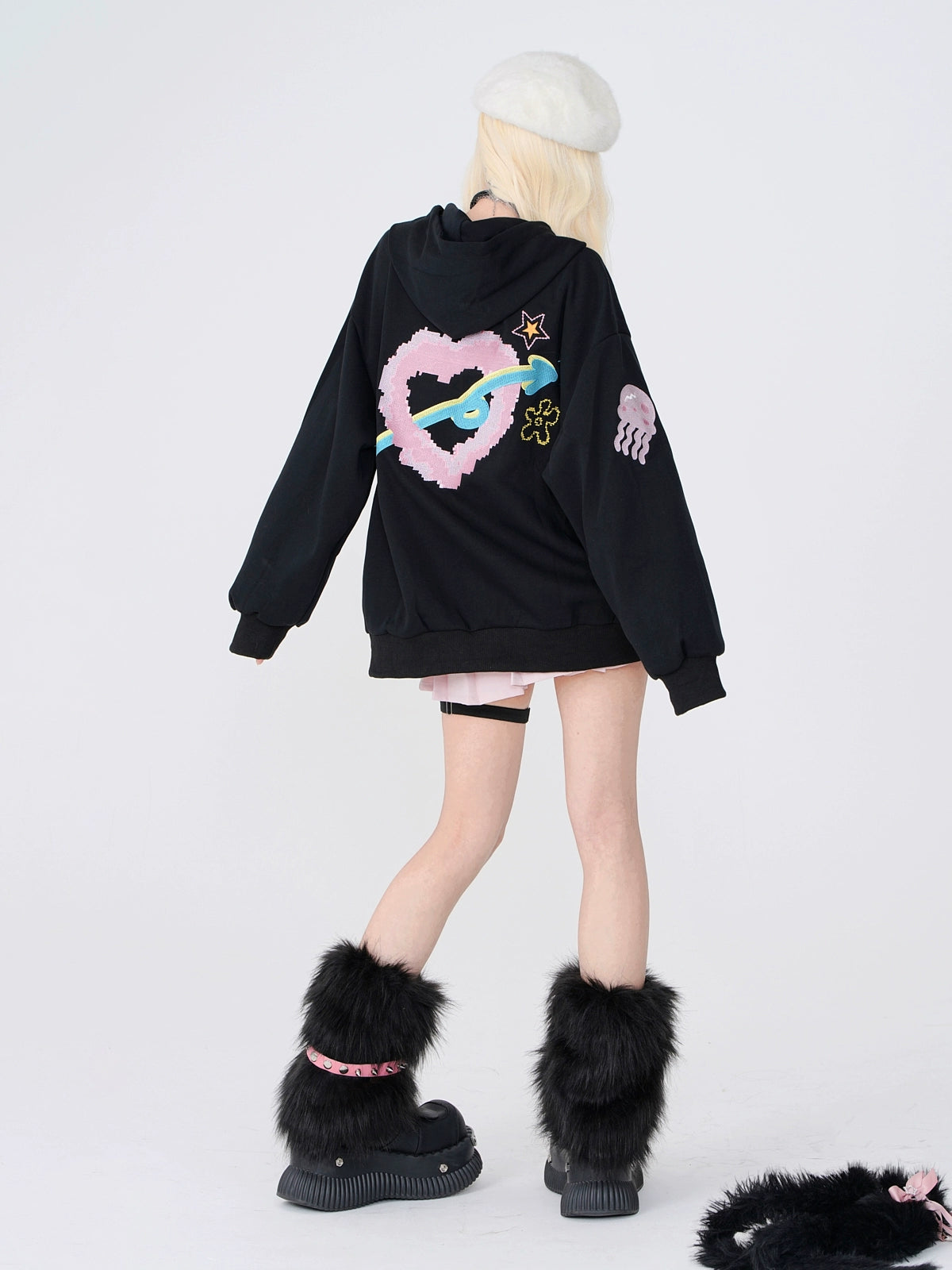 A person with long blonde hair faces away, wearing the Seakoff Kelly Kitty Pastel Kawaii Hoodie featuring adorable embroidered characters. Paired with black furry boots and a white beret, their outfit channels Japanese streetwear against a plain white backdrop.