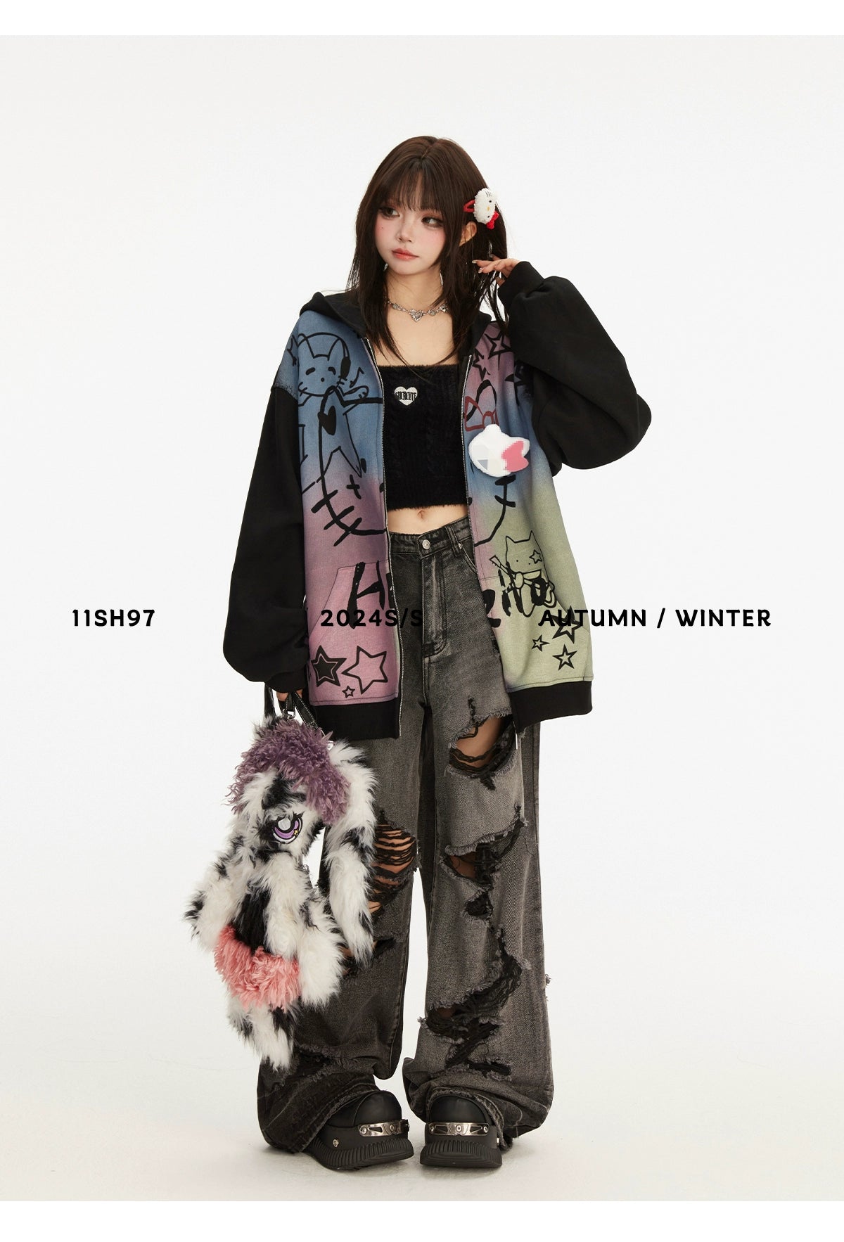 Against a plain background, a person wears an oversized Seakoff Pastel Cat Graffiti Hoodie over a black crop top and distressed wide-leg jeans. They hold a plush accessory, featuring hair styled with a red clip. Text reads &quot;11SH97 2024 S/S AUTUMN/WINTER,&quot; embodying kawaii streetwear vibes.