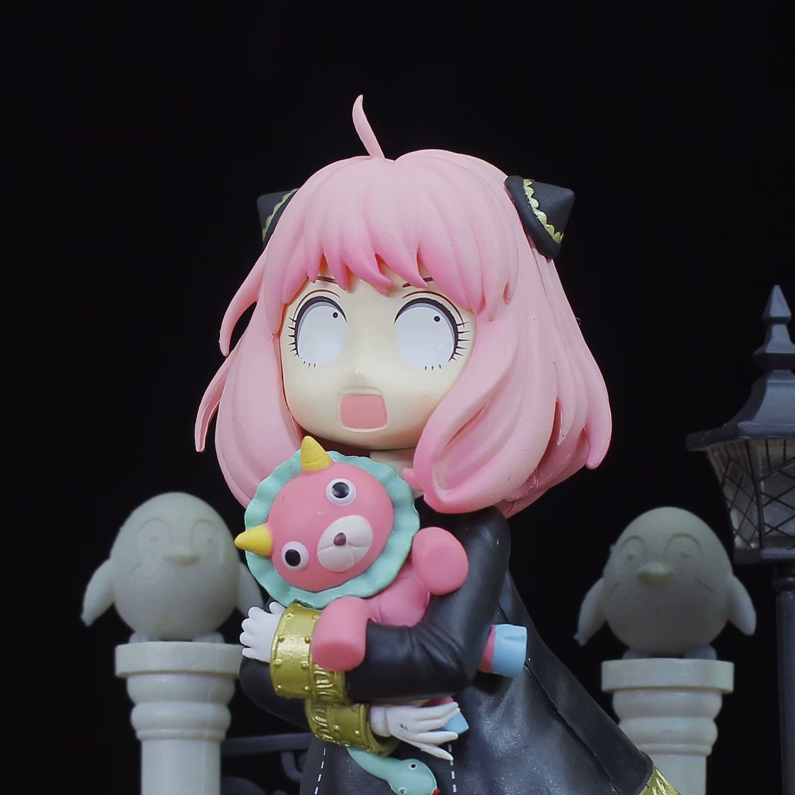 The Spy x Family Anya Forger PVC Figure, a 19 cm cute collectible statue, features Anya with pink hair and black cat-like ears. She holds a plush animal with a horn against stone sculptures and a dark lamppost—a must-have for anime enthusiasts.