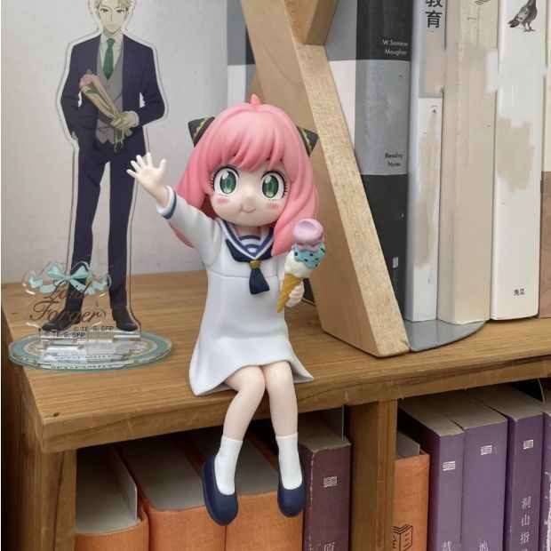 The Spy × Family Anya Forger with Ice Cream Figure, a PVC anime collectible, depicts the pink-haired character in a white sailor-style outfit, holding a pink ice cream cone on a wooden shelf. Behind it is an Anya cutout in a suit and books with various titles.