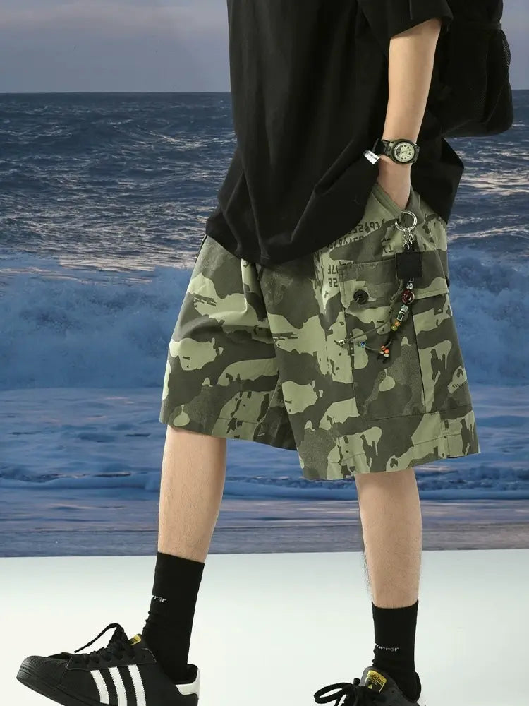 A person in a black shirt, black socks, black Adidas sneakers, and Men's Green Camo Cargo Shorts with Drawstring Waist and Button Flap Pockets stands by the ocean with waves in the background.