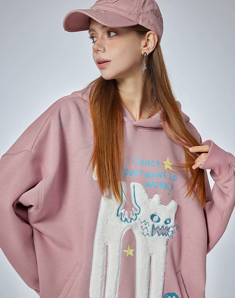 A young woman with long red hair wears Seakoff's "Oh No! I Don’t Want to Work!" oversized graphic hoodie, featuring a lazy cat design. She gazes left, left hand in her hair, right hand in the pocket, embodying casual streetwear vibes with a pink cap.