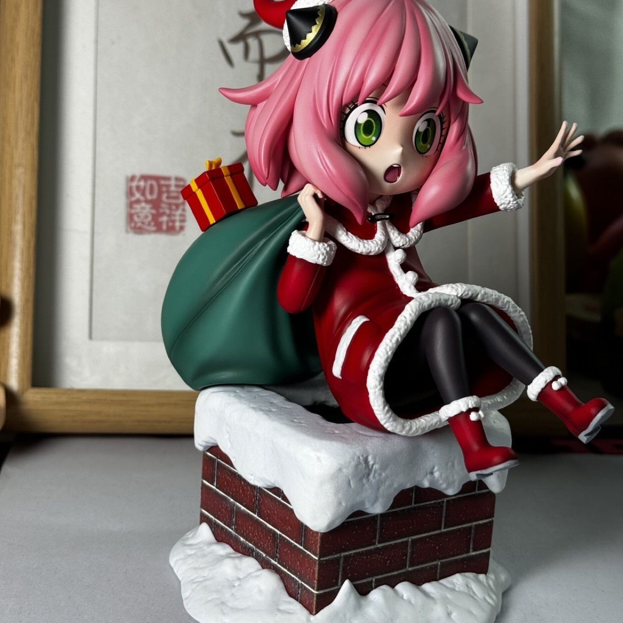 The Anya Forger Christmas Outfit Figure - PVC Anime Collectible from Spy × Family is perched on a snow-covered chimney. Featuring signature pink hair and red-and-white winter attire, she holds a green sack with a surprised expression, enhanced by blurred framed artwork—ideal for anime collectors.