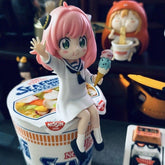 Anya Forger in a white sailor dress with pink hair sits on a large Nissin cup noodle holding ice cream, while another figure in an orange hat enjoys noodles. This scene is part of the "Anya Forger with Ice Cream Figure" from the Spy × Family PVC Anime Collectible series.