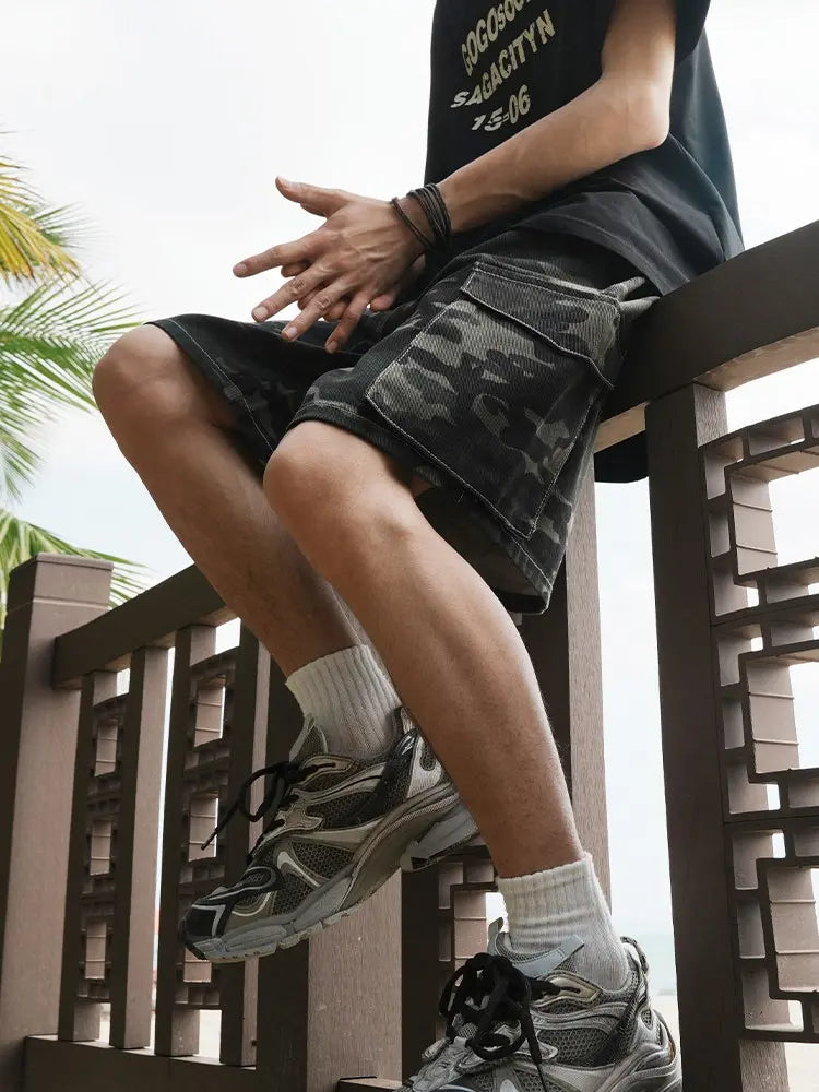 A person, showing only their lower body and hands, sits casually on a railing outdoors with palm leaves in the background, wearing Men's Baggy Camo Cargo Shorts with Elastic Drawstring Waist and Flap Pockets, along with a black T-shirt, white socks, and gray sneakers.