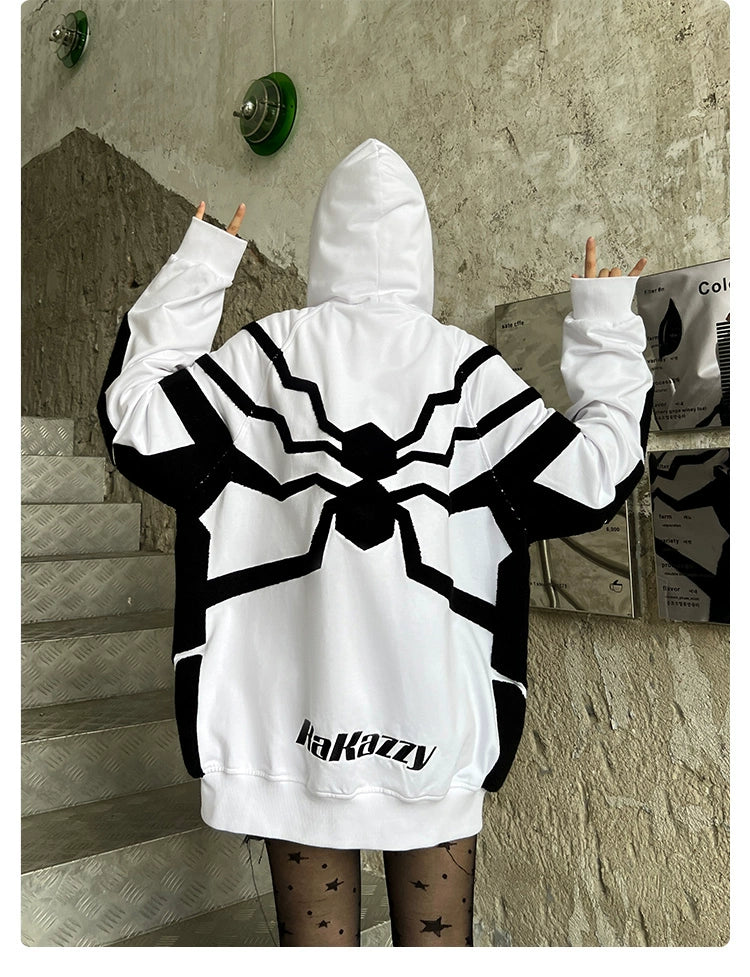 A person wears a Seakoff Venom-Inspired White Hoodie with a black geometric spider emblem on the back, hood up, and hands in a peace sign. Made from 400GSM cotton and featuring &quot;Hakazzy&quot; at the bottom, it stands against a textured wall and metal stairs backdrop.