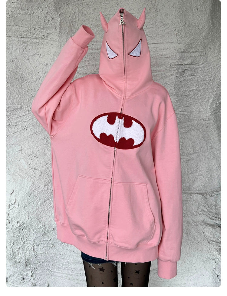 A person wears a Seakoff Batman-Inspired Hoodie—a 400GSM oversized pink zip-up superhero sweatshirt with bat-like ears and embroidered eyes on the fully zipped hood. The chest displays a red and white emblem like bat wings, set against a textured white wall—a must-have for superhero fans.