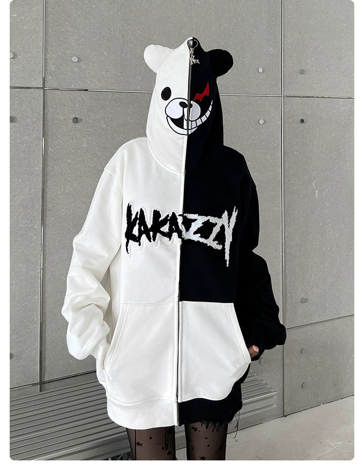 A person is wearing a Seakoff Monokuma-Inspired Hoodie, an oversized black and white zip-up Danganronpa sweatshirt, with &quot;KAKAZZY&quot; text on the front. The gray tiled wall behind enhances the bold look, paired with black patterned tights.