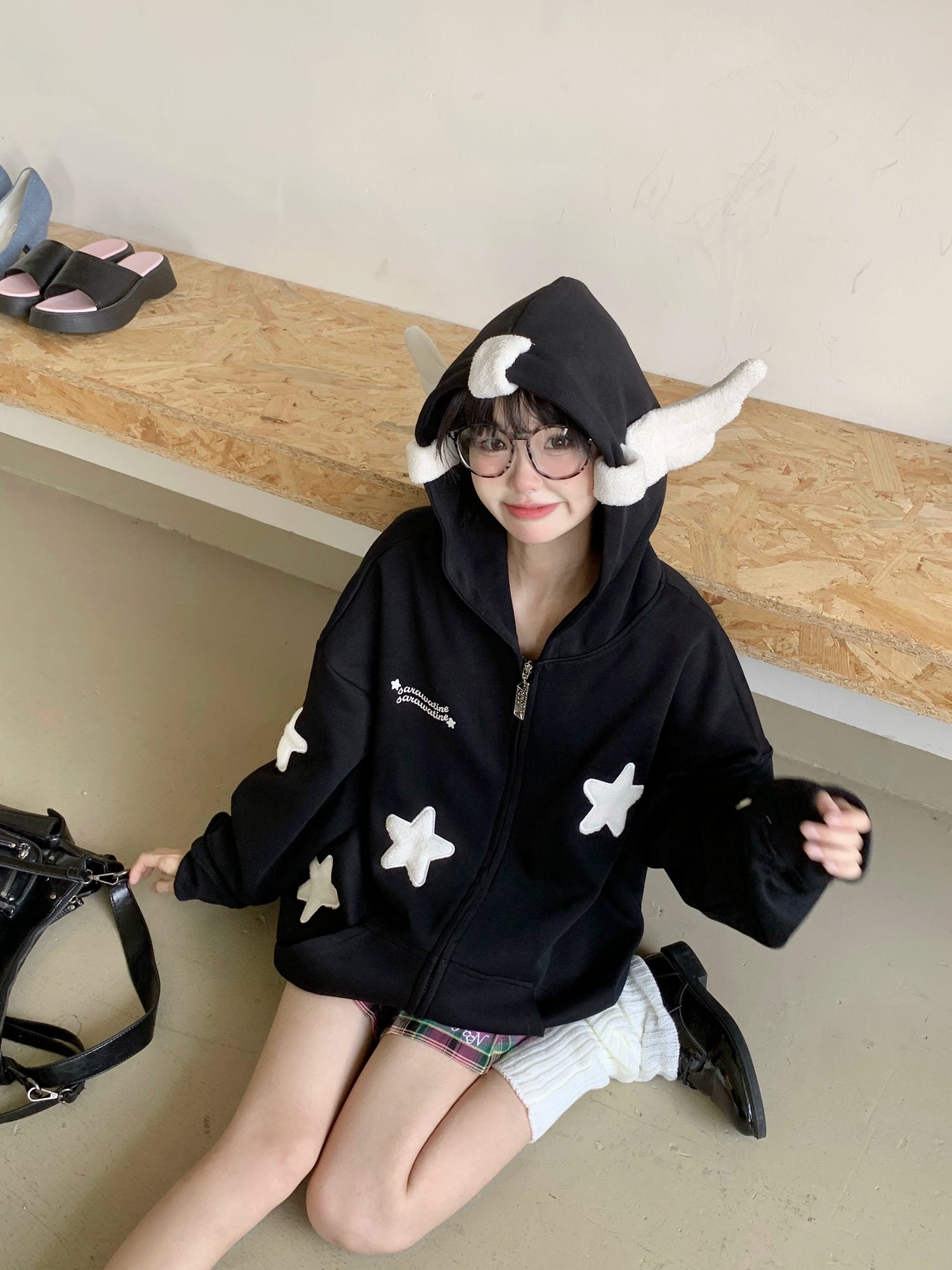 Wearing a Seakoff Kawaii Star Elf Hoodie, featuring 3D ears and soft star appliques, a person sits on the floor with glasses, smiling, while a black handbag and shoes rest nearby, embracing whimsical fashion.