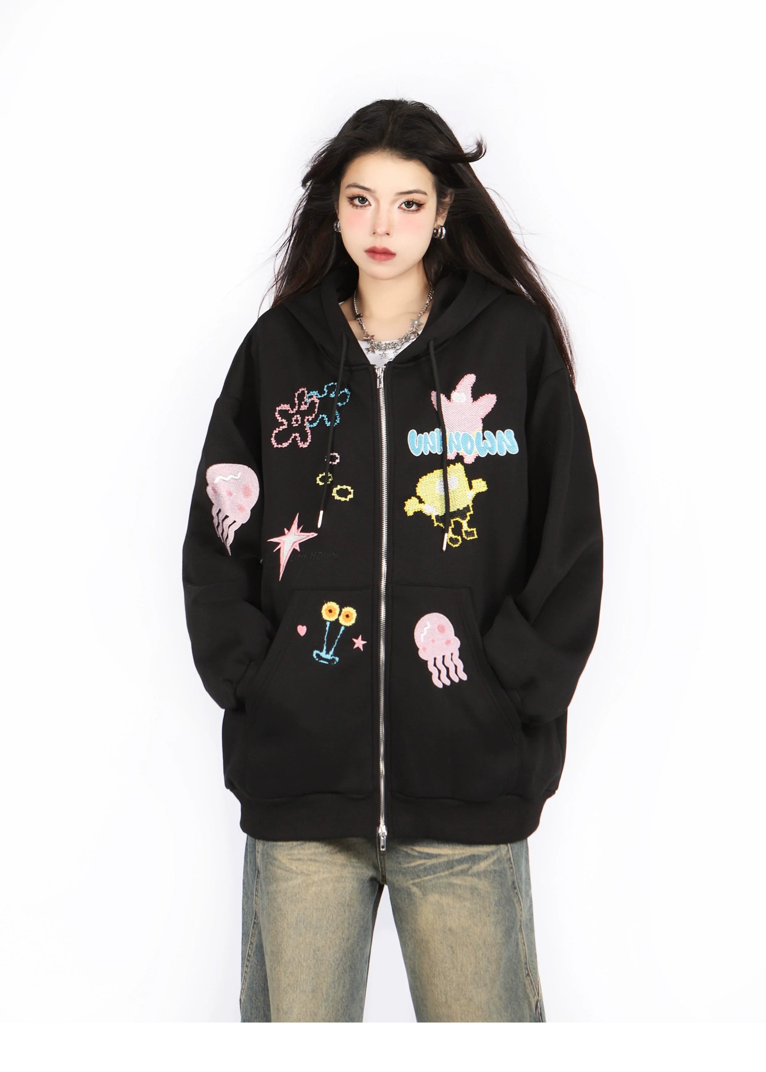 Against a white backdrop, someone wears the Seakoff Kawaii Pastel Cartoon Hoodie, showcasing playful embroidered designs of jellyfish, flowers, and abstract shapes that echo anime fashion. This oversized black hoodie perfectly complements loose-fitting jeans for a playful yet stylish look.