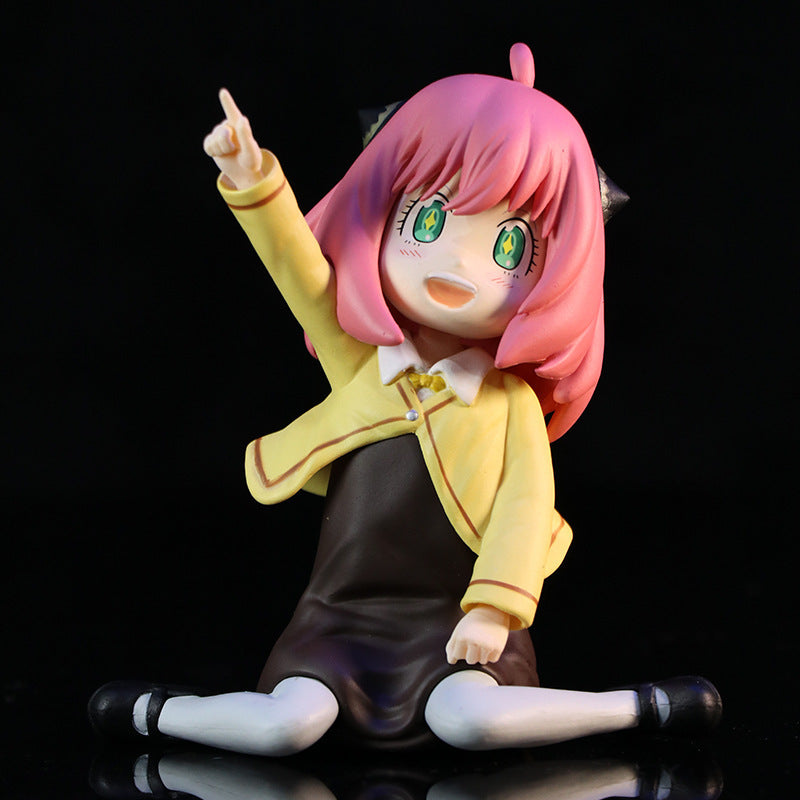 The Anya Forger figure, a Spy × Family collectible, depicts the beloved animated character with pink hair and star-shaped eyes in a yellow school uniform, seated on the ground with one arm raised. This PVC anime figure includes an interchangeable face, making it essential for anime fans.