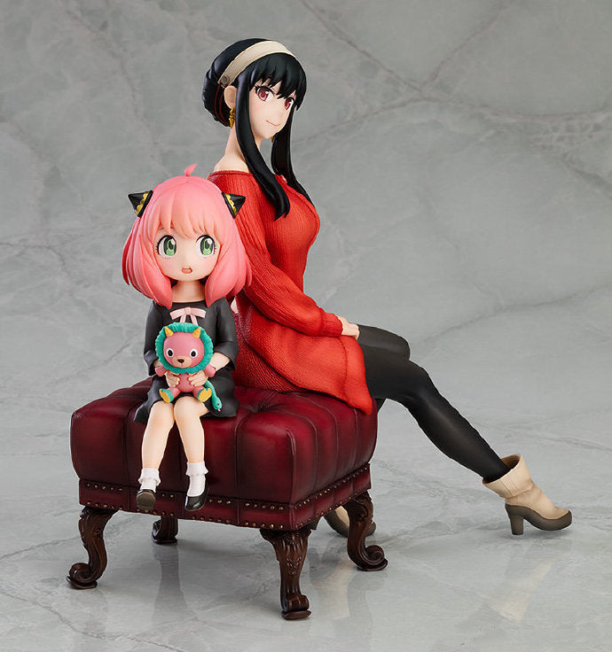 Two anime figures, Anya and Yor from Spy × Family, sit on a red cushioned stool in a 15 cm PVC figure set. Yor is in a red dress with crossed legs, while Anya has pink hair and a cat-ear headband, holding a toy. This charming display celebrates their mother-daughter bond against a light gray background.