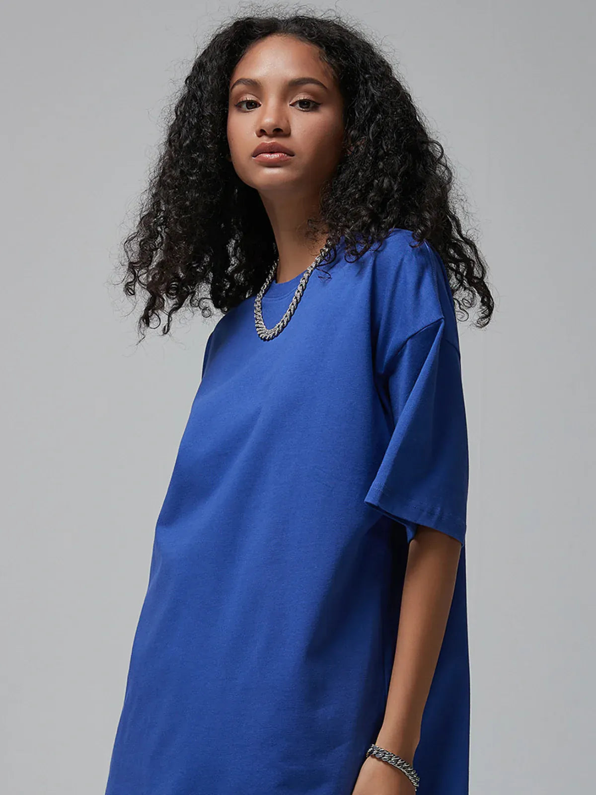 A woman with curly hair wears the Custom Anime Character Royal Blue T-Shirt—personalized, made from 230GSM 100% cotton—paired with a silver chain necklace and bracelet, standing against a plain gray background.