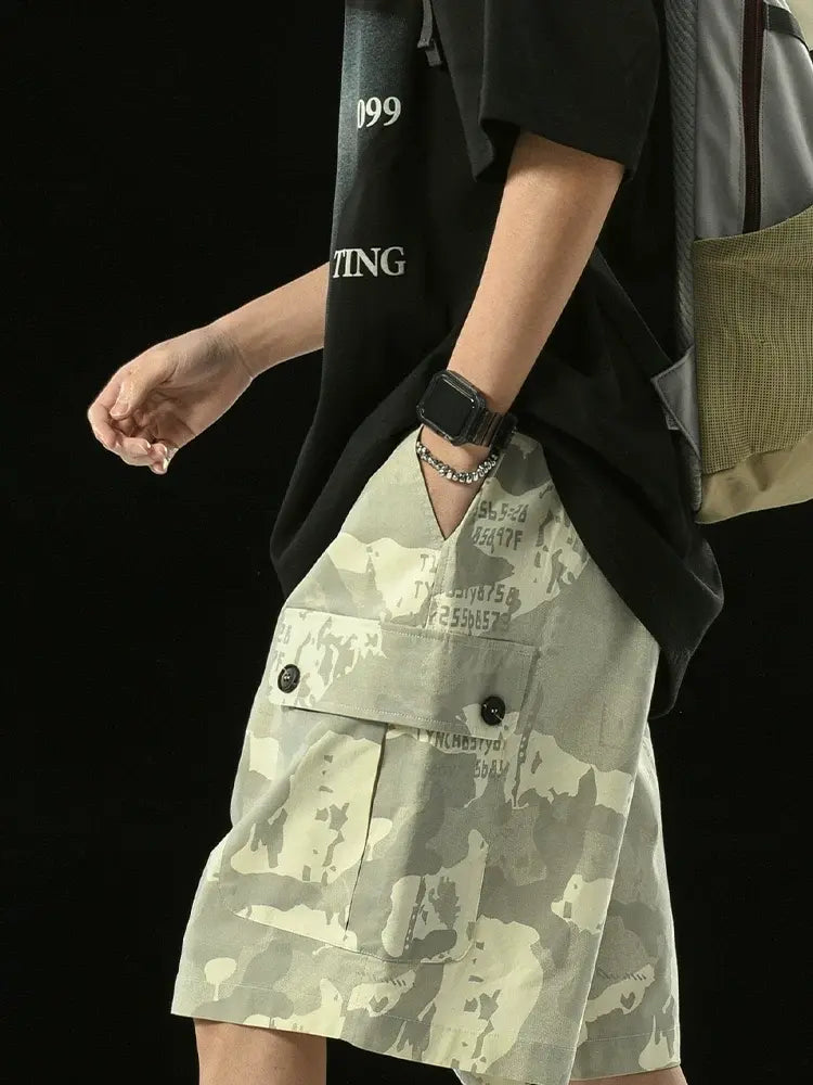 A person stands against a black background wearing a black t-shirt, smartwatch, gray backpack, and Men's Grey Camo Cargo Shorts with Button Flap Pockets and Elastic Waist; accessories are visible and their hand is in their pocket.