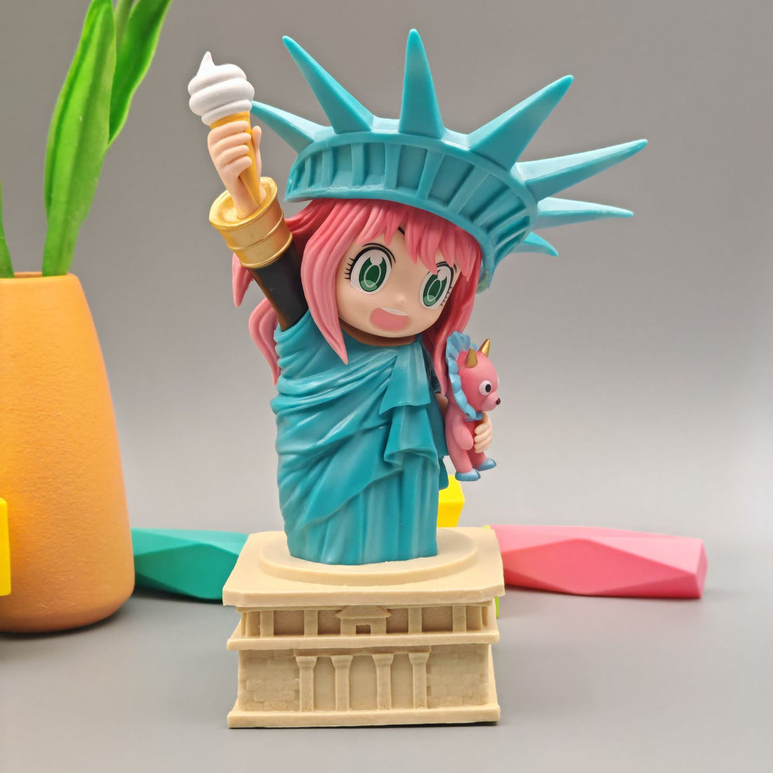 The Spy x Family Anya Forger Statue of Liberty PVC Figure features a cute chibi style with pink hair, holding an ice cream and paired with a small pink sidekick. This 18.5 cm figure by Spy × Family is framed with a potted plant and vibrant accents, capturing whimsical charm.