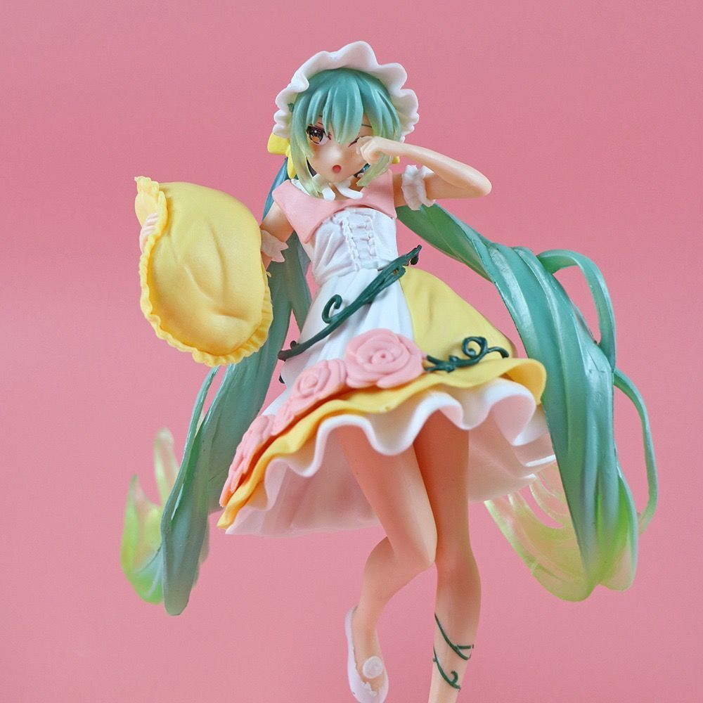 The Hatsune Miku Sleeping Beauty PVC Figure, standing at 20cm, captures Miku with long green hair and a yellow floral dress with a white hat. She holds a yellow pillow and is set against a pink backdrop, appearing mid-step in this adorable collectible with box.