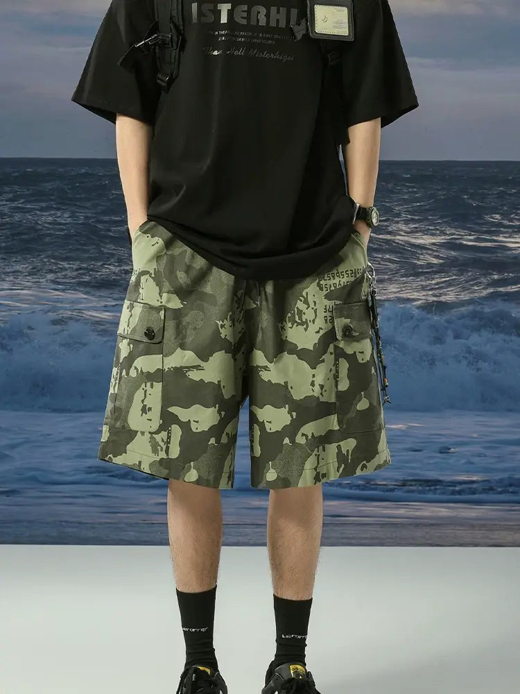 A person stands with hands in the pockets of Men's Green Camo Cargo Shorts with Drawstring Waist and Button Flap Pockets, paired with a black T-shirt, black socks, and black shoes, against an ocean waves backdrop.
