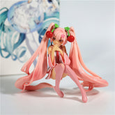 The Hatsune Miku Cherry Sitting Pose 11cm PVC Figure is a charming collectible, featuring the character with long pink twin tails and red spheres. She wears a pink outfit with knee-high socks in a playful sitting pose, set against a colorful blurred background.