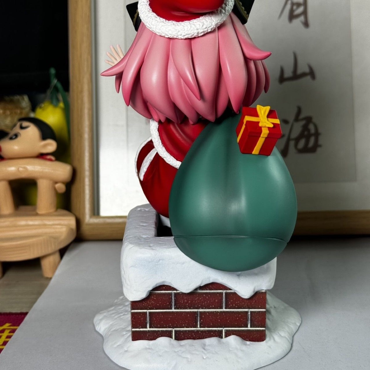 The Anya Forger Christmas Outfit Figure by Spy × Family features a pink-haired Santa on a snowy brick chimney, holding a green sack and a gift. A plush toy and framed calligraphy complete the background, making it perfect for anime collectors.