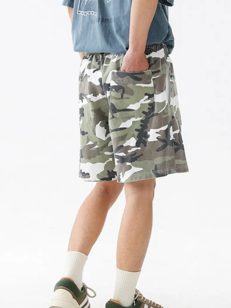 A person wearing Men's Woodland Camo Cotton Drawstring Shorts in green, brown, and white, along with a blue t-shirt, white socks, and green sneakers stands with one hand in their pocket against a plain white background.