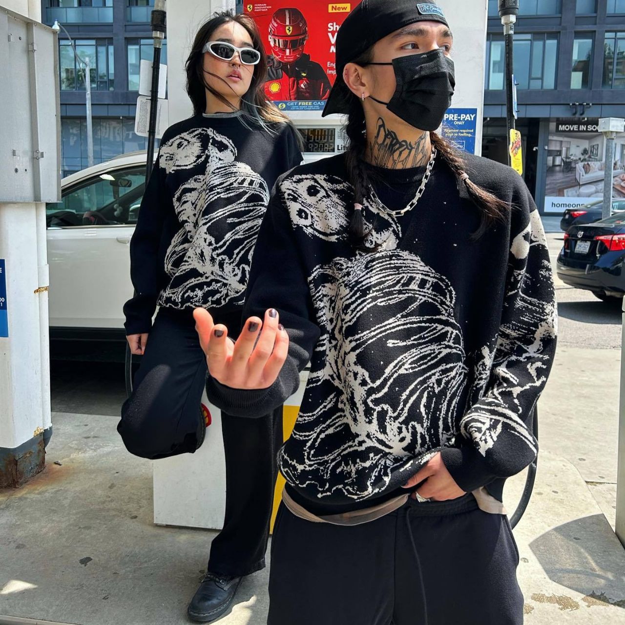 Two people at a gas station wear matching Seakoff's Premium Skeleton Ribcage Knit Sweaters – Gothic Streetwear Jacquard Design. One, in sunglasses, holds a drink while the other gestures with a black mask. Their striking alternative fashion stands out amid the cars and buildings.