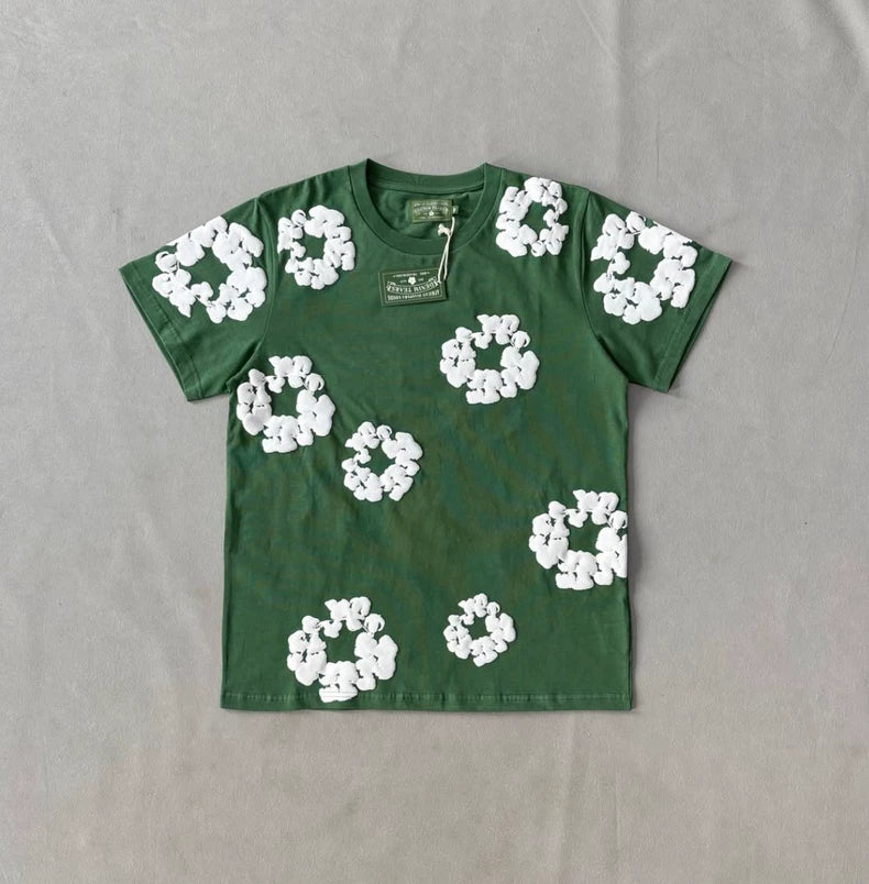 The Denim Tears Green Cotton Wreath Allover Print T-Shirt features a relaxed oversized fit and white wreath-like designs across the front, shown flat on a light gray surface with a tag attached to the collar.