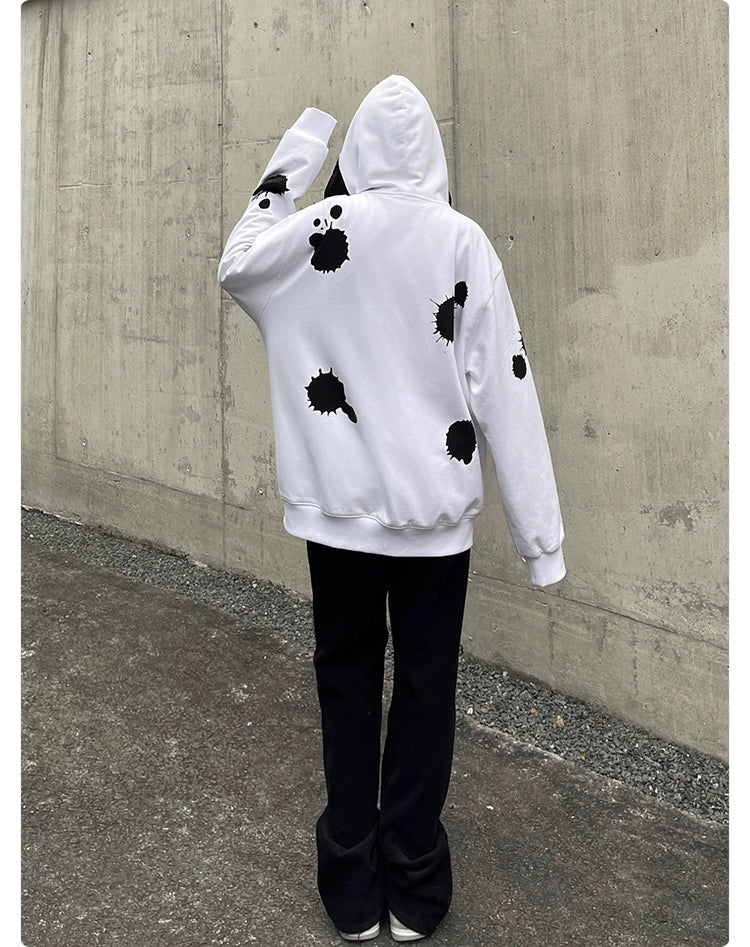 A person in a Seakoff Monochrome Ink Splatter Hoodie, 400GSM oversized white and black zip-up streetwear sweatshirt, paired with black pants, poses against a gray concrete wall with the hood up and one arm raised, capturing an edgy streetwear vibe.