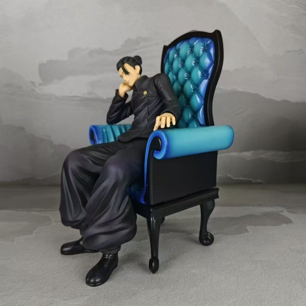 The Jujutsu Kaisen Satoru Gojo &amp; Suguru Geto Sitting Posture PVC Figure Set features a 24 cm statue of a character in an ornate chair with blue upholstery, dressed in black clothing, exuding a relaxed vibe with one arm on the armrest and the other near their face.