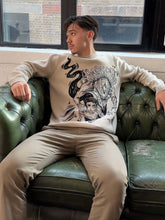 A man sits on a green leather sofa by a window, wearing the Premium Yuta Okkotsu-Inspired Knit Sweater from Jujutsu Kaisen, featuring a cursed spirit jacquard design. He pairs this with beige pants and relaxes with an arm draped over the back of the sofa.