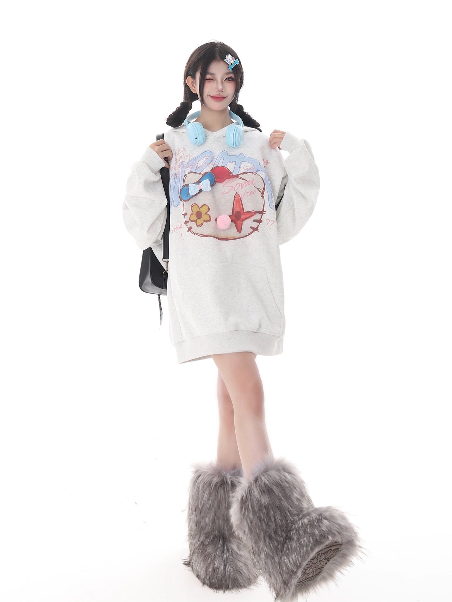 Against a white background, a person showcases streetwear flair with Seakoff’s Kawaii Pom-Pom Crown Hoodie, featuring a unique cat graphic and 3D pom details. They wear furry boots, blue headphones, sport pigtails, carry a black backpack, and add kawaii touches with playful accessories.