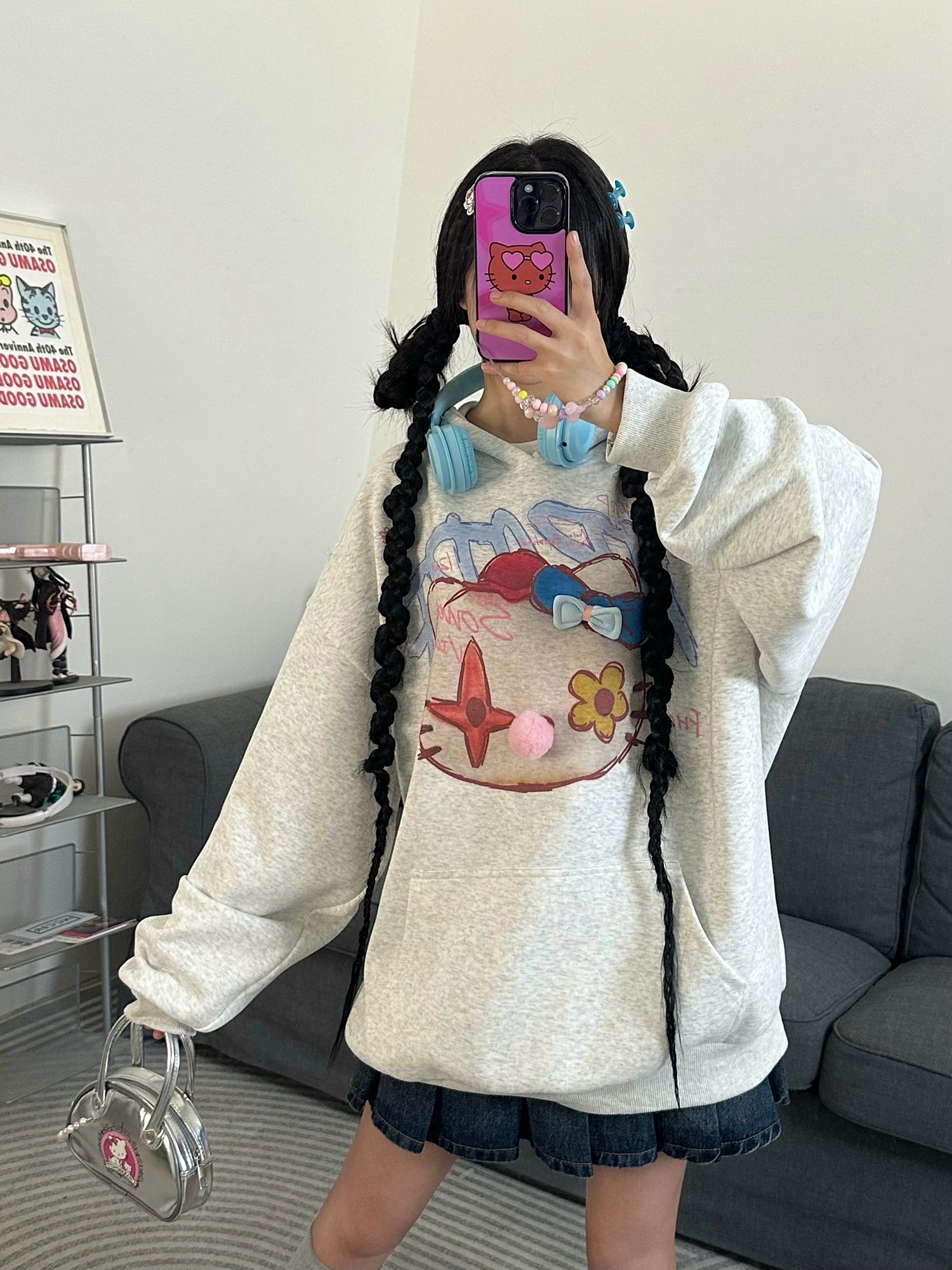 In a living room, a person snaps a selfie in streetwear: the Seakoff Kawaii Pom-Pom Crown Hoodie featuring a unique cat graphic and 3D pom details paired with a denim skirt. Blue headphones frame their face, their phone case shows a cartoon bear, and they hold a small silver bag enhancing the kawaii aesthetic.