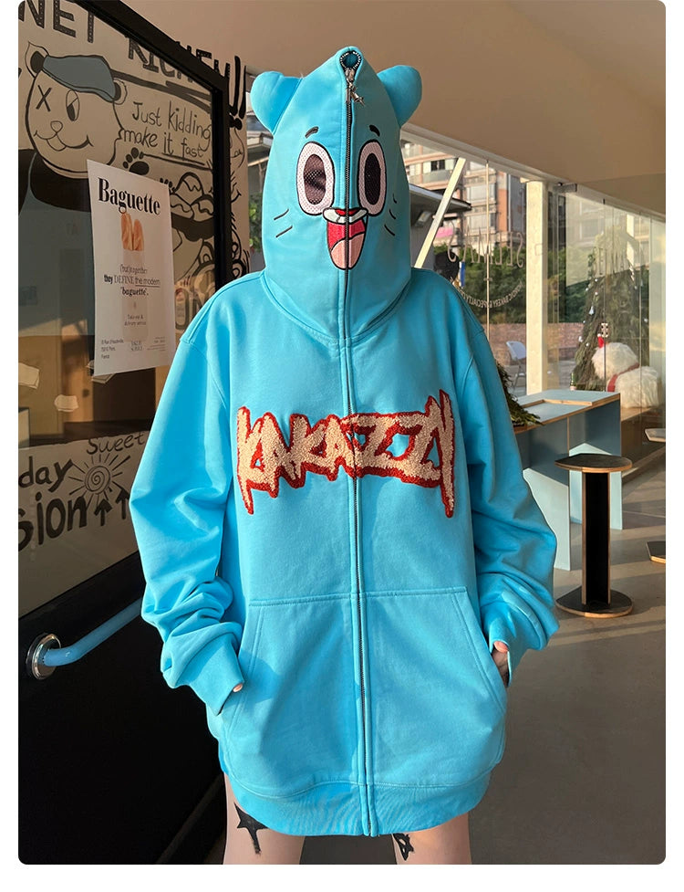 Indoors against a modern backdrop with a window, someone wears the Seakoff Adorable Blue Cartoon Hoodie, made from 400GSM cotton featuring cartoonish ears and a playful face design, complemented by cartoon-style text on the front.