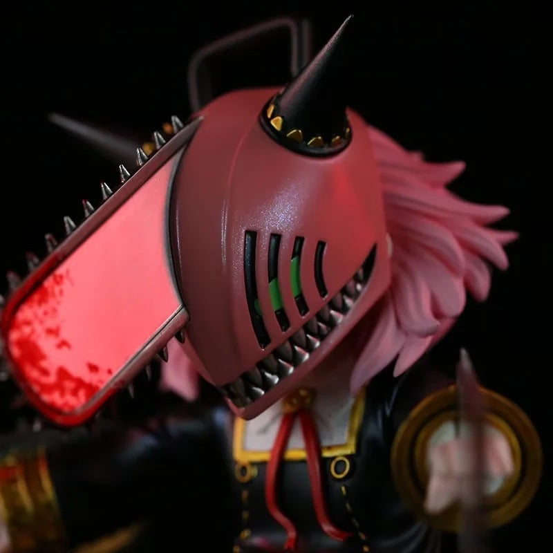 A close-up of the Chainsaw Man Anya Forger PVC Figure from Spy × Family shows chainsaw-themed headgear with serrated teeth, a pink mane, black horns, and green eyes through slits. The 17.5 cm collectible statue wears a detailed outfit with a striking red tie.