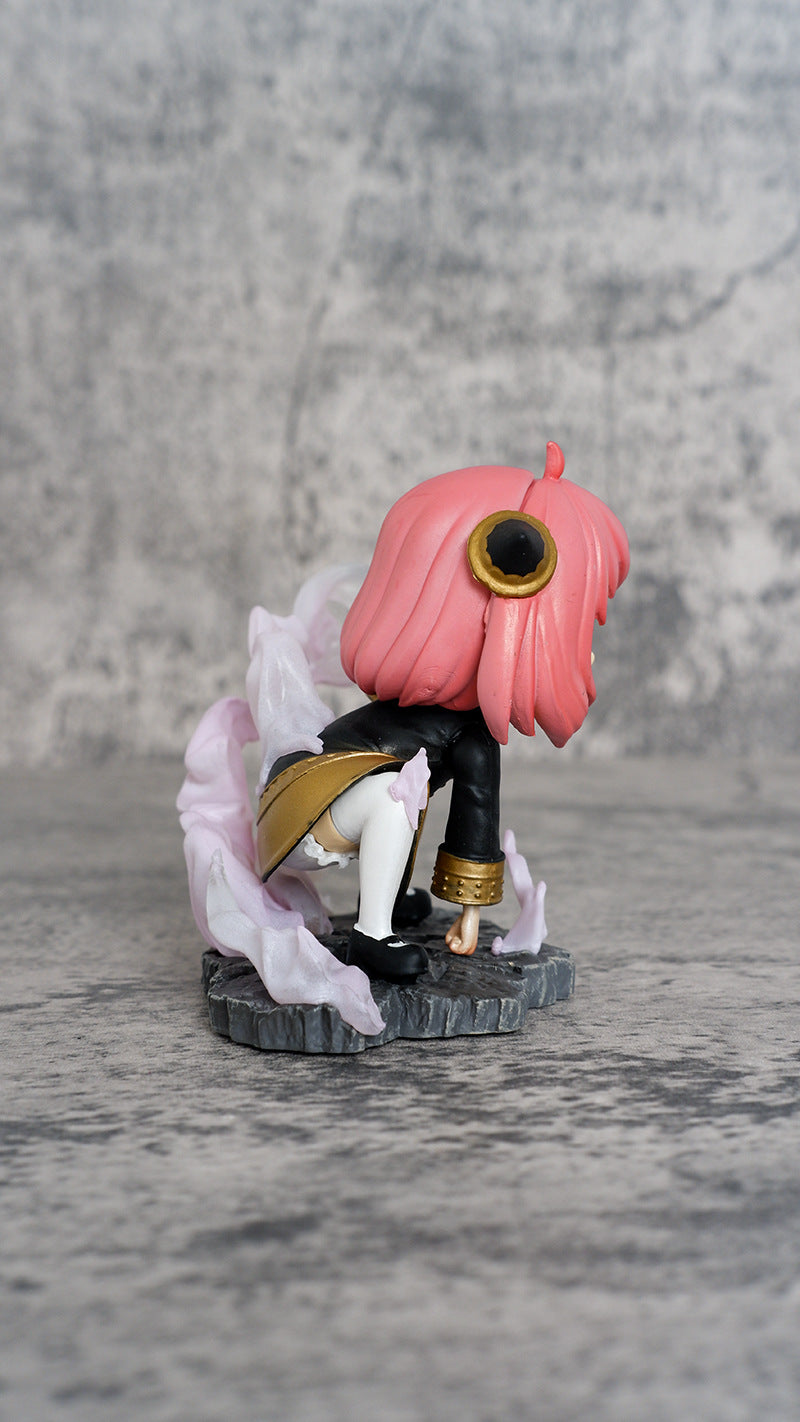 The Spy × Family Anya Forger PVC Figure depicts an anime character with pink hair and a unique circular eye, dressed in black and gold on a textured gray base with translucent accents. Included is a double head replacement, with a mottled gray backdrop.