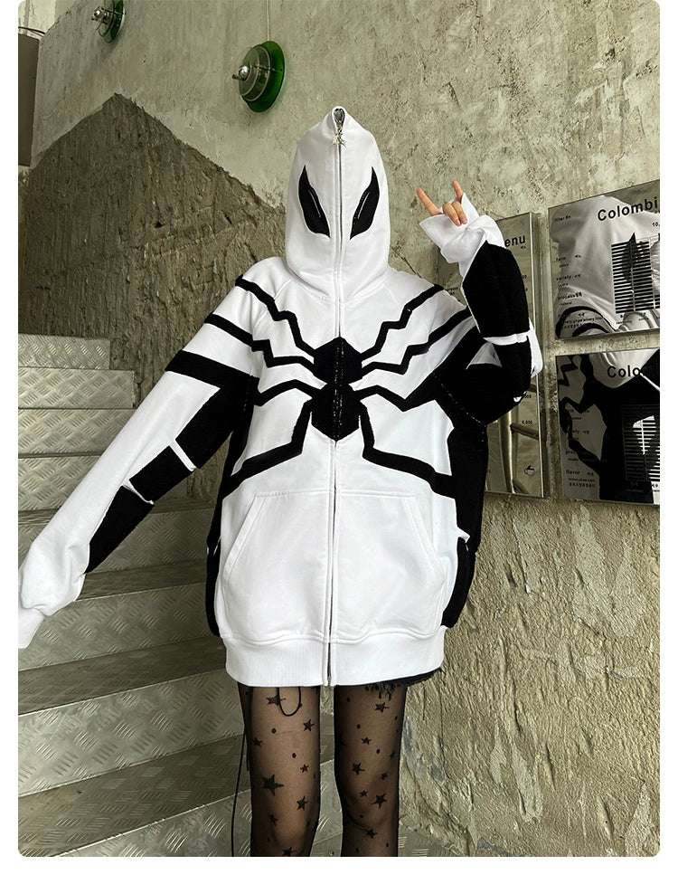 Standing on stairs making a peace sign, a person wears the Seakoff Venom-Inspired White Hoodie, an oversized zip-up superhero sweatshirt featuring spider web and eye designs made from 400GSM cotton. Star-patterned tights add flair against textured concrete walls adorned with posters.