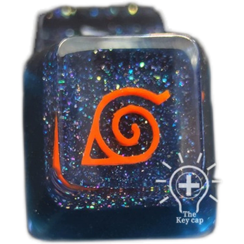 A close-up of the Naruto Symbols Keycap from the Hidden Village, Uchiha, Akatsuki set features a glossy, glittery blue background with an orange leaf symbol. Encased in high-quality resin for an illuminated effect, &quot;The Keycap&quot; logo is placed at the bottom right.