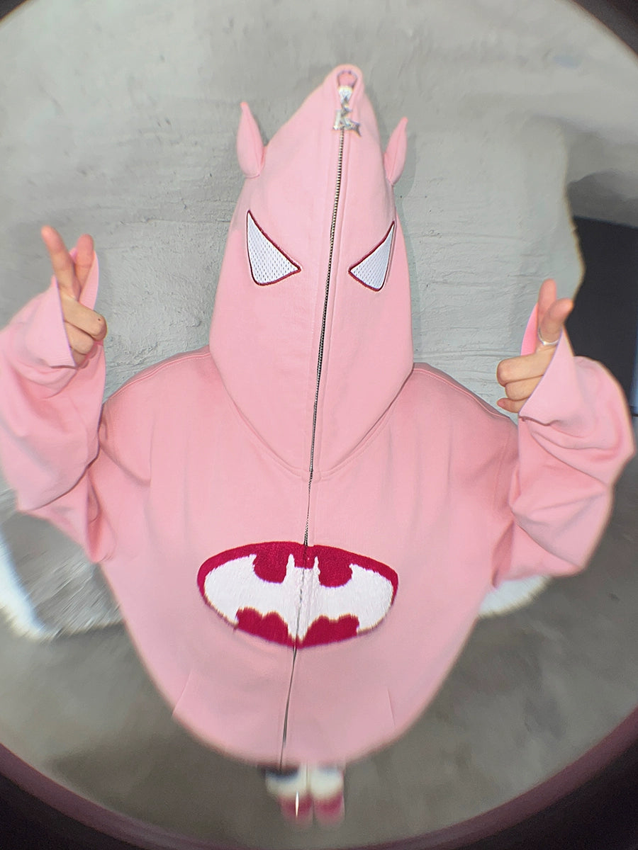 A person wears the Seakoff Batman-Inspired Hoodie, a pink 400GSM oversized zip-up sweatshirt that covers their face except for mesh eye cutouts. It features a superhero logo on the front. With peace signs in both hands, this look is perfect for superhero fans.