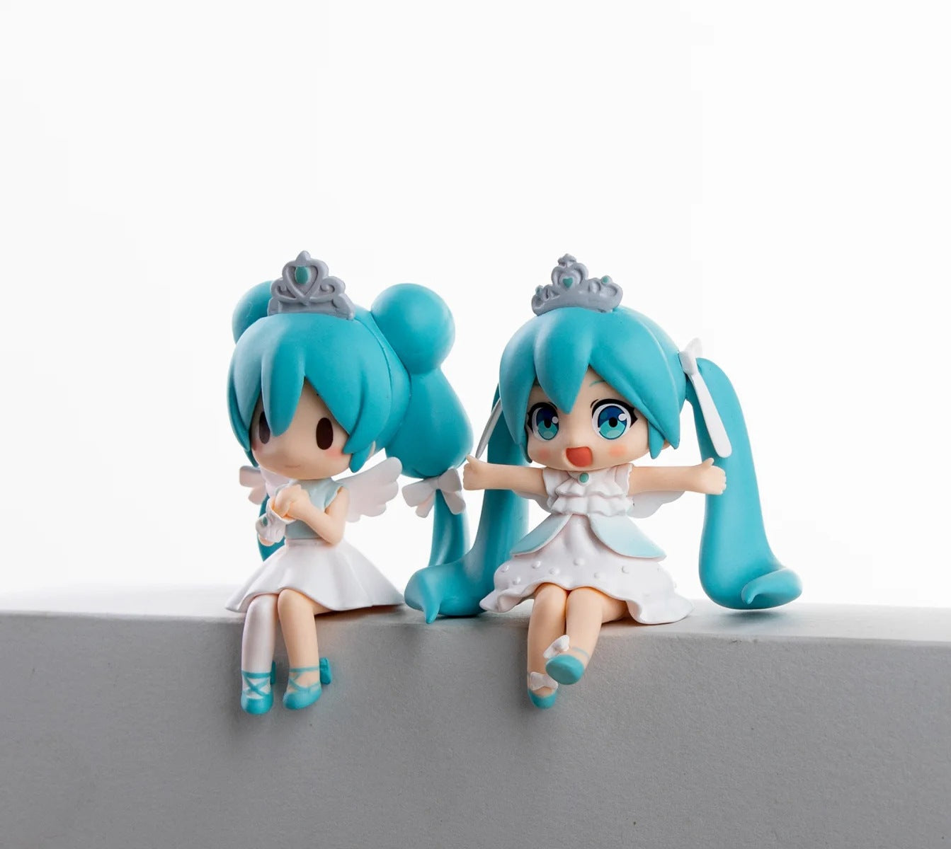 The Hatsune Miku Prayer Hugging Angel Princess 2-piece Set features two 7.5cm PVC figures with blue hair and an Angel Princess aura. Dressed in white, crowned, and wing-adorned, one sits serenely while the other poses with arms outstretched on a gray ledge.