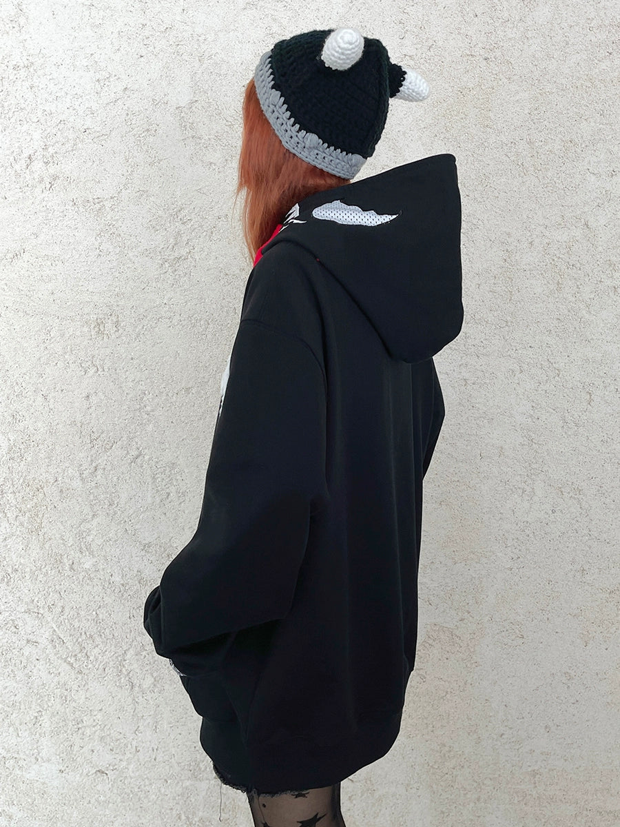 A person with long hair, wearing a Seakoff Venom-inspired hoodie featuring a signature tongue and spider logo design, along with a knit hat with white pom-poms, stands against a light textured wall, facing away from the camera.