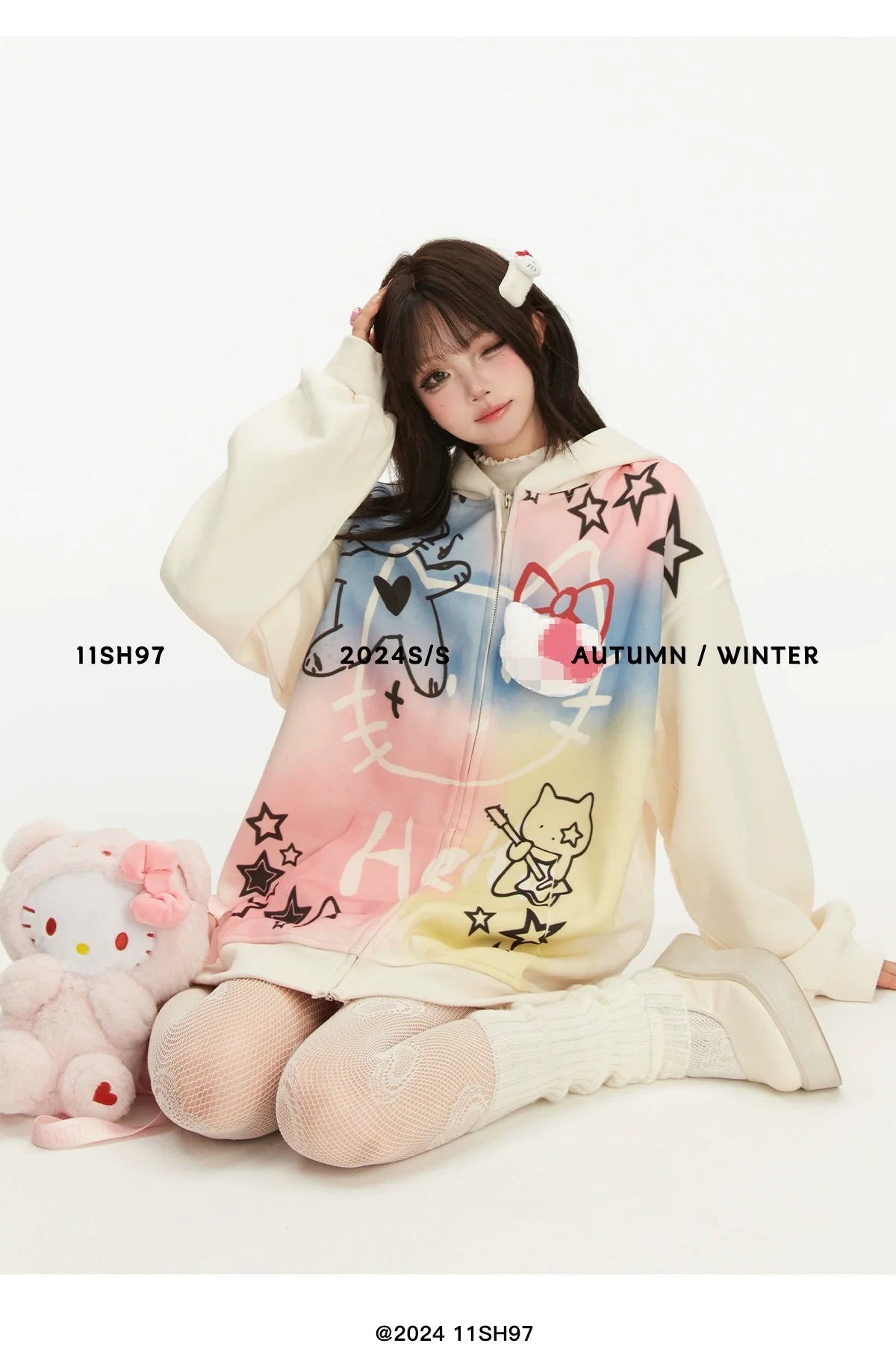 A person sits on the floor in a Seakoff oversized Pastel Cat Graffiti Hoodie, embracing their knees. They're surrounded by a plush toy and have a hair clip in their hair. The plain white background features text overlays promoting Seakoff's kawaii streetwear collection for 2024.