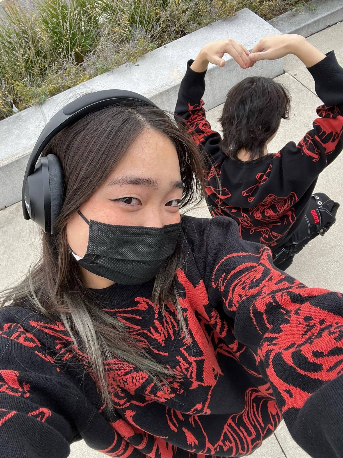 Two individuals wear matching Jujutsu Kaisen knit sweaters from the Yuji Itadori & Nobara Kugisaki Jacquard Design. The foreground figure, resembling Yuji with headphones, takes a selfie, while the background figure mimics Nobara's style and sits forming a heart shape with their arms.