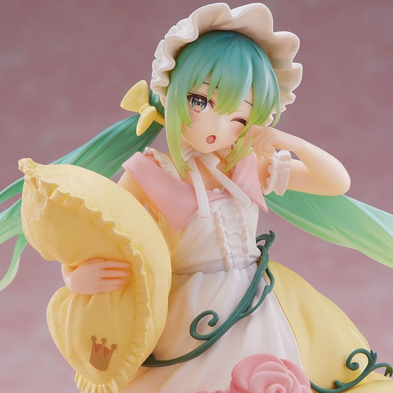 This adorable 20cm Hatsune Miku Sleeping Beauty PVC Figure showcases the iconic character in a frilly white and pink dress with a bonnet, holding a yellow pillow with an emblem, winking sweetly against a blurred pink background. Comes as an adorable collectible with box.