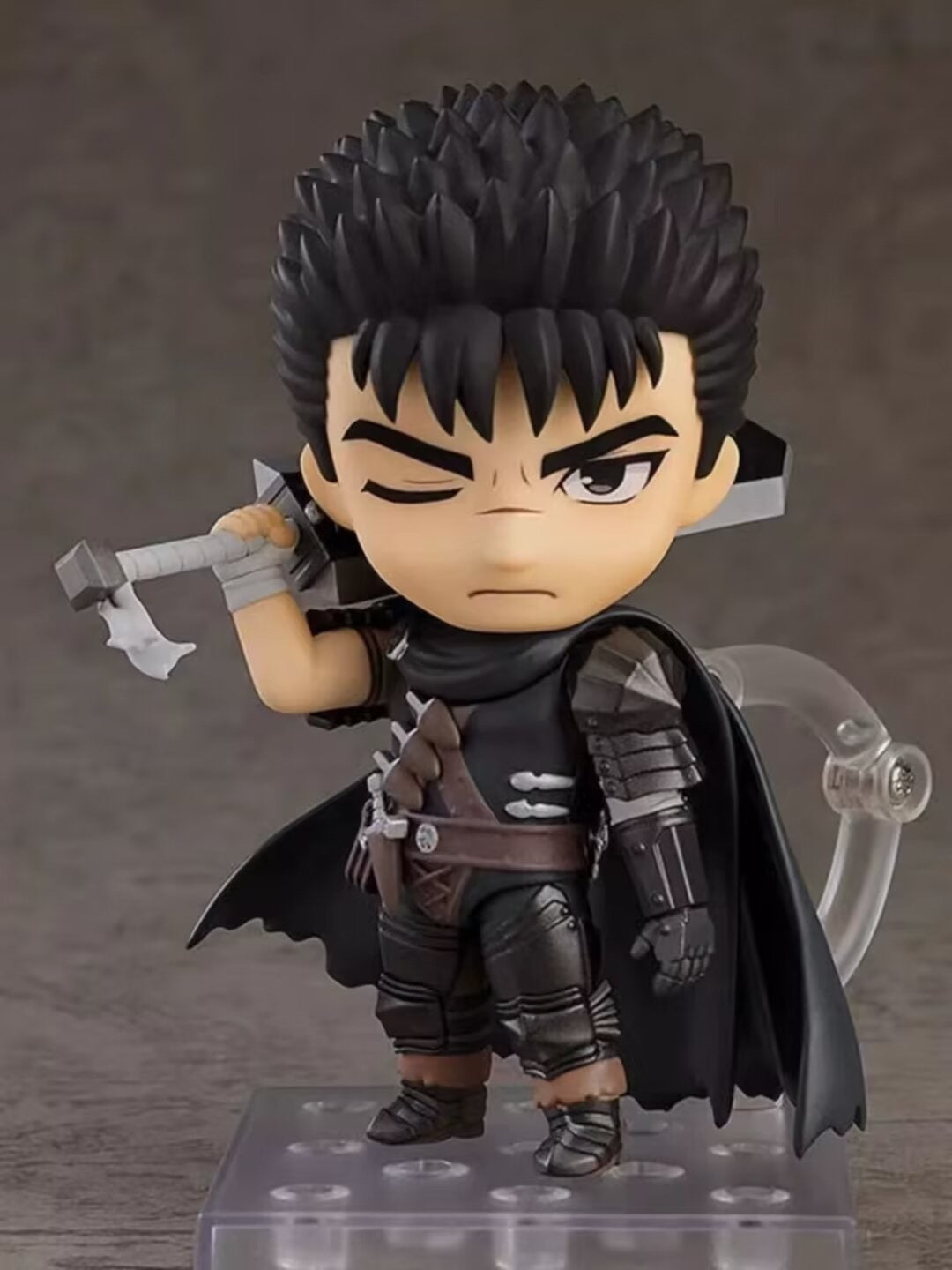 The Berserk Guts Nendoroid Action Figure is a chibi-style PVC figurine featuring spiky black hair, a stern look with one eye closed, and movable joints. It dons a black cloak and armor while holding a sword on its shoulder, standing on a clear base—perfect for any anime figure collection.