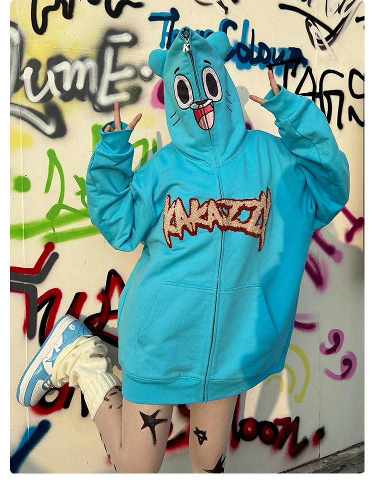 Wearing Seakoff’s Adorable Blue Cartoon Hoodie, a person strikes peace signs in front of a graffiti wall. The oversized 400GSM cotton hoodie with playful face design matches their shoes and the colorful backdrop perfectly.