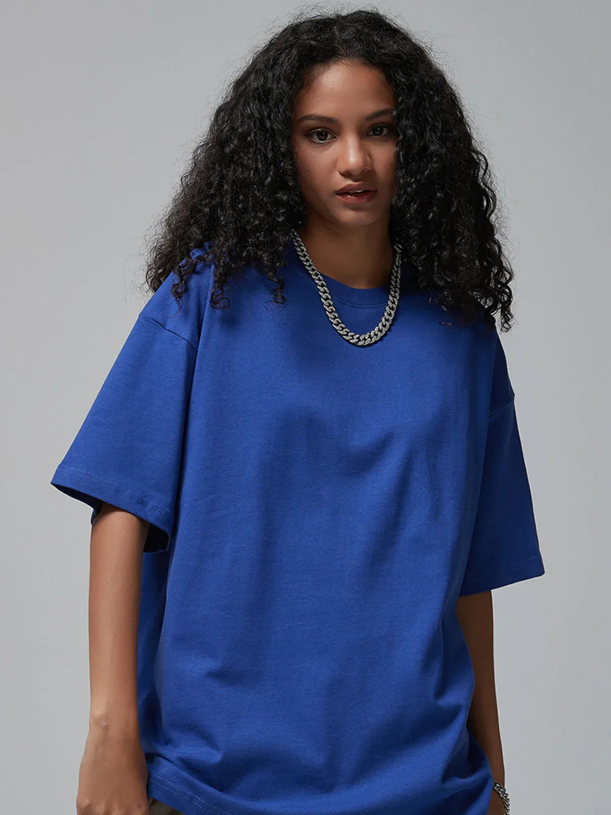 A person with long curly hair wears the Custom Anime Character Royal Blue T-Shirt - Personalized 230GSM 100% Cotton Tee and a silver chain necklace, standing against a plain gray background.