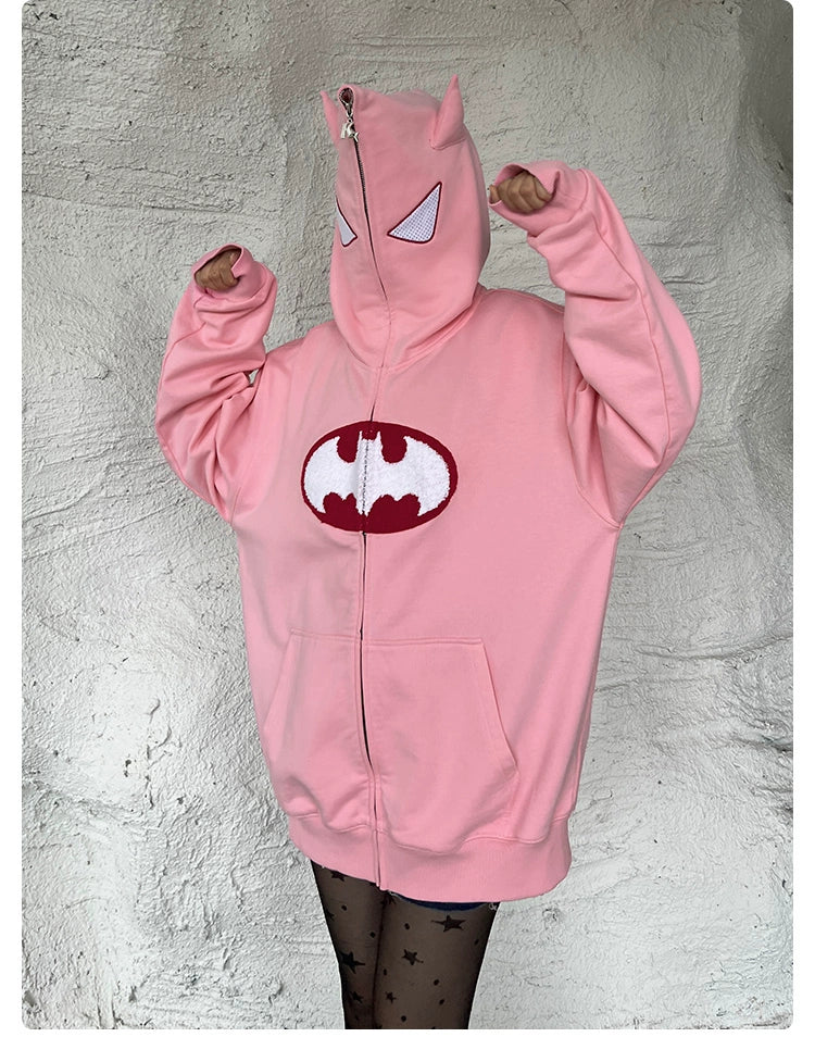 A person wears the Seakoff Batman-Inspired Hoodie, a 400GSM oversized pink zip-up sweatshirt with white eye designs and cat ears. Featuring a bat symbol on the front, it’s ideal for superhero fans. Paired with star-patterned leggings, they strike a playful pose with raised hands.