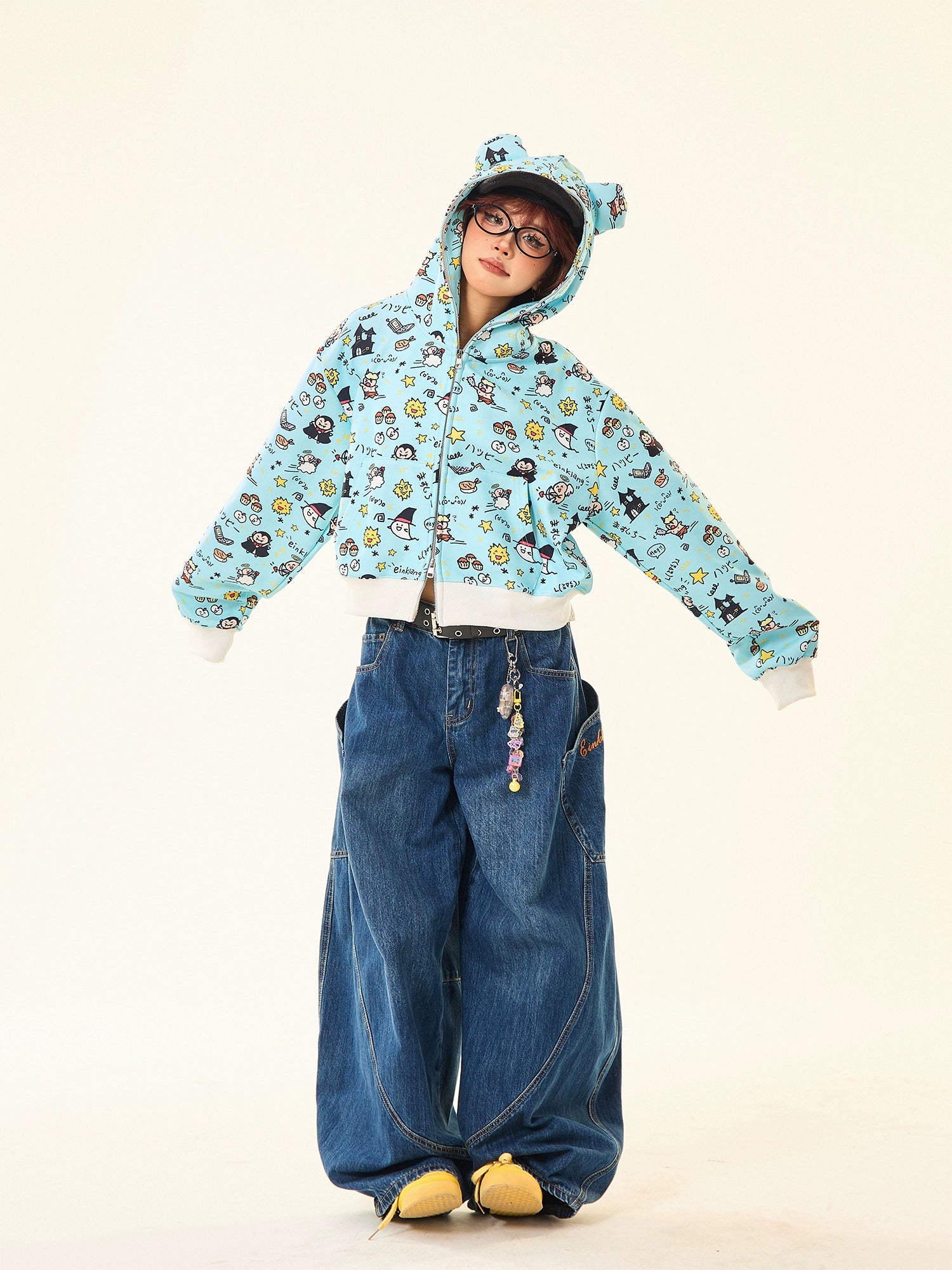 A person in a whimsical streetwear look wears the Seakoff Kawaii Cartoon Bear Hoodie, featuring cute ears and a fun blue print. Paired with oversized blue jeans, yellow shoes, and glasses, they stand against a neutral background with a keychain hanging from their jeans.